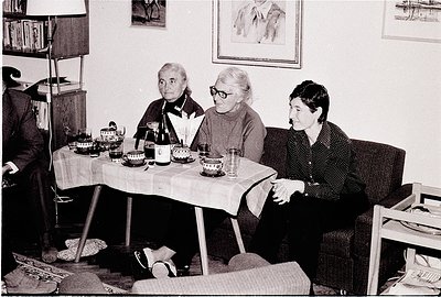 Three women seated at a table in a vintage indoor setting, likely mid-20th century. Glasses, bottles, and plates suggest a ca...