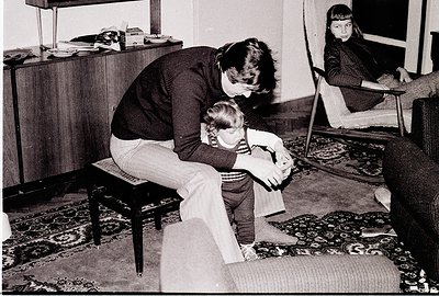 A tender 1970s domestic scene: an adult kneels beside a young child on a wooden stool, offering comfort. A second child sits ...