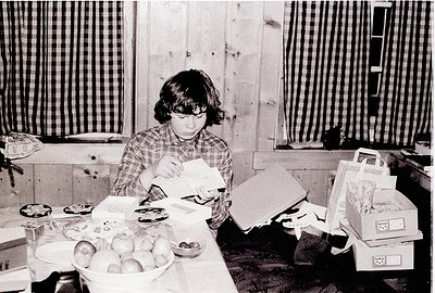 A young person in a plaid shirt organizes paper packages on a cluttered wooden table surrounded by household items. Fresh fru...