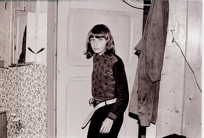 A young girl in a patterned sweater stands in a dimly lit indoor space, holding a belt. The wallpaper features floral pattern...