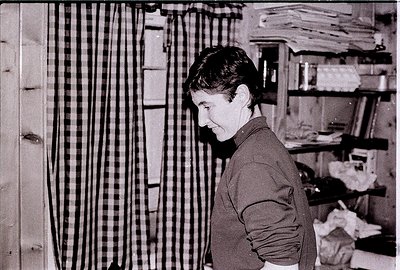 Black-and-white portrait of a person standing in a textile workshop, surrounded by stacked fabric rolls and woven textiles. T...