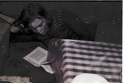 Vintage black-and-white photo of a child resting on a striped mattress, reading an open book. Patterned blanket and a round p...
