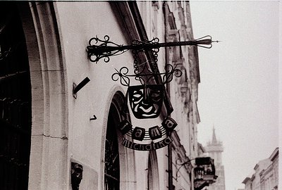 Black-and-white street sign featuring a stylized, grinning mask with decorative wreath elements, mounted on ornate metal brac...