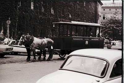 Vintage urban scene featuring a horse-drawn tram labeled "Towarzystwo" alongside mid-century cars. The tram’s vintage design ...