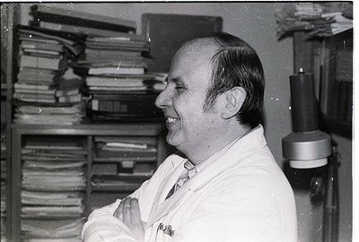 Mid-century academic in lab coat surrounded by stacked binders, likely a university or research setting. Classic 1960s–1970s ...