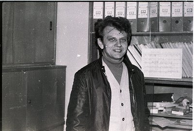 Black-and-white office portrait of a man in a leather jacket and button-down shirt, standing beside metal filing cabinets and...
