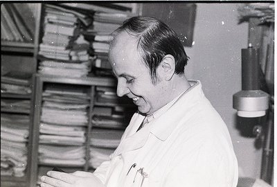 Mid-20th century black-and-white photo of a man in a lab coat examining rolled microfilm or film reels in a storage cabinet. ...