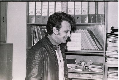 Mid-century office setting featuring a man in a leather jacket and collared shirt, smiling at the camera. Surrounding him are...