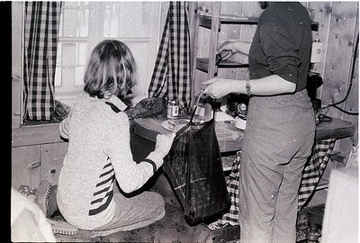 Two women in a mid-century kitchen, likely 1960s–1970s, preparing food. One kneels, holding a jar, while the other stands, po...