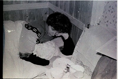 Black-and-white photo of a person sleeping on a mattress in a damaged, partially collapsed room. Exposed bricks and debris su...