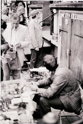 Vintage black-and-white photo of three men engaged in detailed craftwork in a dimly lit workshop. One man sits at a table wit...