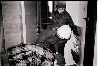 Mid-20th century indoor scene: A child in winter attire (hat, coat, gloves) carries a large white fur coat over their shoulde...