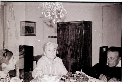 Vintage black-and-white interior shot of two elderly individuals seated at a table adorned with floral arrangements. The woma...
