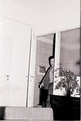 Mid-century indoor portrait: person in doorway holding a framed photo, framed mirror above, floral-patterned wallpaper. Likel...