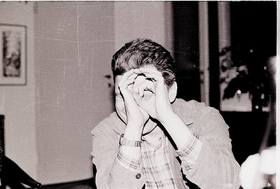 Black-and-white candid of an individual covering their face with hands, seated indoors. Wearing a checkered shirt with a wris...
