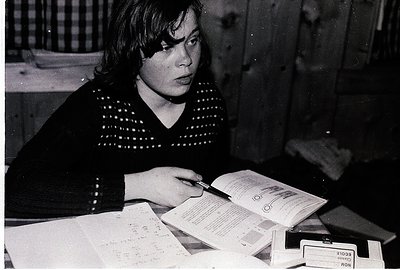 Vintage black-and-white photo of a woman in a patterned sweater studying a passport and handwritten notes, likely at a desk. ...
