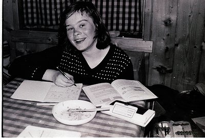 Young girl writing at a wooden table in a rustic indoor setting, likely mid-20th century. She wears a polka-dot sweater and h...
