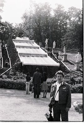 Mid-20th century black-and-white photo of a man in a military-style jacket posing in a grand park setting. Behind him, a symm...