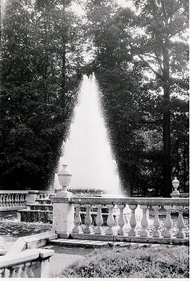 Classic 19th-century fountain with symmetrical stone balustrade and tall, conical water jet framed by mature trees. Likely Eu...
