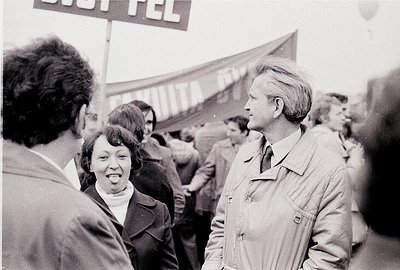 Black-and-white protest scene featuring two central figures in mid-conversation, surrounded by a crowd. The man wears a light...