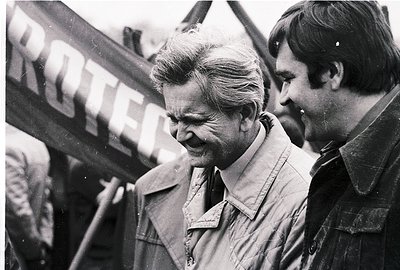 Black-and-white photo of two men at a public event, likely 1960s–1970s. The man in focus wears a light jacket and appears eng...