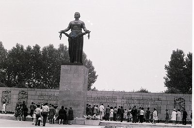 Monumental statue of a robed figure holding a scroll, standing atop a stone pedestal with inscribed plaques. Crowd gathers in...