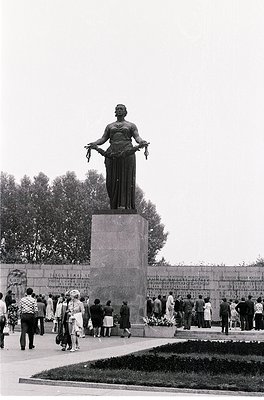 Monumental statue of a robed woman holding a wreath atop a stone pedestal, surrounded by engraved plaques. Crowd of people in...