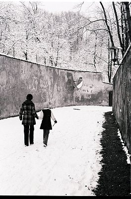 Two figures walk hand-in-hand along a snow-covered path beside a high concrete wall, framed by bare winter trees. The woman w...