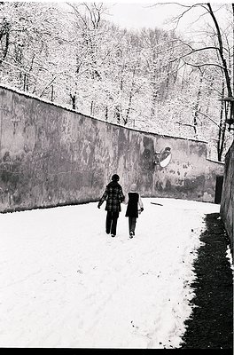 Two children walk hand-in-hand on a snow-covered path beside a concrete wall, framed by bare winter trees. Mid-20th century u...