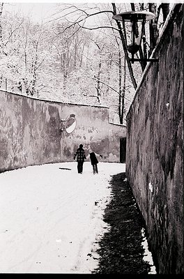 Black-and-white street scene featuring two figures walking along a snow-covered alley flanked by high walls. Snow accumulates...