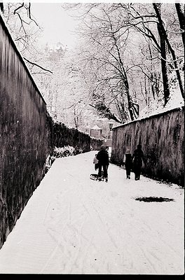 Black-and-white street scene featuring three children sledding down a snow-covered path flanked by high concrete walls. Bare ...