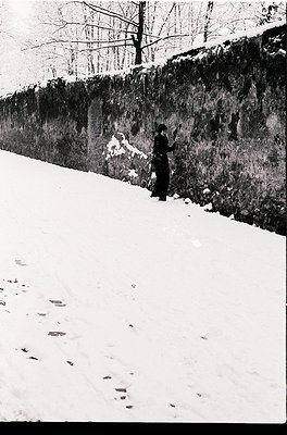 Black-and-white street scene featuring a lone figure in winter attire—long coat, hat, and gloves—standing beside a stone wall...