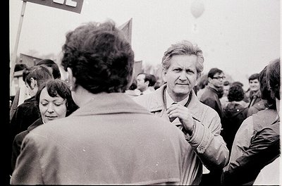 Black-and-white street protest with dense crowd, 1970s–1980s Eastern Bloc. Central figure in jacket gestures toward camera, s...