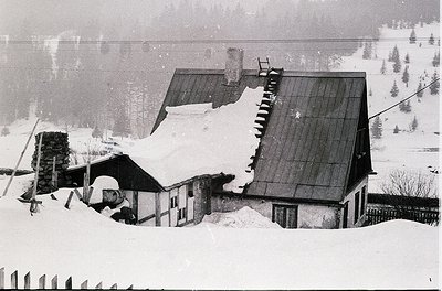 Snow-covered alpine cabin with steeply pitched roof, partially buried in winter. Rustic stone chimney and wooden exterior sug...