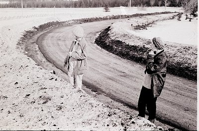 Mid-20th century black-and-white snapshot of two figures on a snow-covered rural road, likely . Adult holds child’s hands, bo...