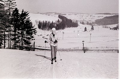 Mid-century cross-country skier in snowy forest, posing with poles. Classic 1960s winter attire—long-sleeve top, gloves, and ...