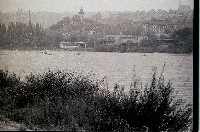 Vintage black-and-white riverside scene with dense foliage in foreground. Mid-20th century urban landscape featuring low-rise...
