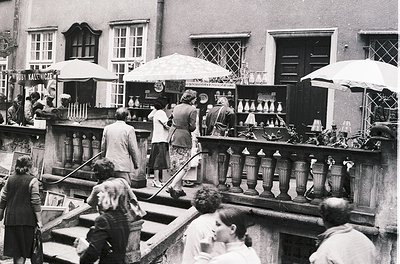 Vintage black-and-white courtyard market featuring brassware stalls under umbrellas. Crowd browsing ornate pitchers, teapots,...
