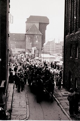 Crowded urban procession in a narrow alleyway, flanked by brick buildings and industrial architecture. A hearse carries a fla...
