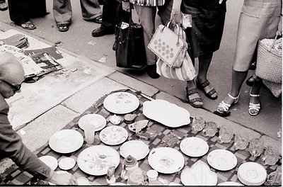 Street market scene featuring handmade ceramic plates and bowls displayed on a pavement table. Mid-20th century style with vi...