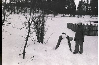 Two individuals build a snowman in a wooded area, mid-20th century. One kneels shaping snow, the other stands holding a cylin...