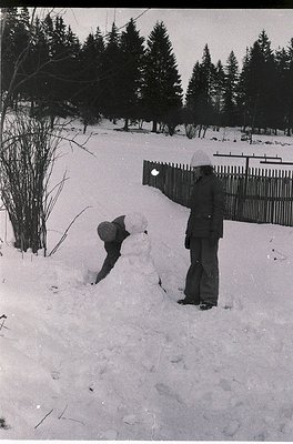 Black-and-white snapshot of two children building a snowman in a residential backyard, surrounded by snow-covered trees and a...