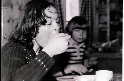 Black-and-white snapshot of two children drinking from cups at a café table, likely mid-20th century. The girl in the foregro...