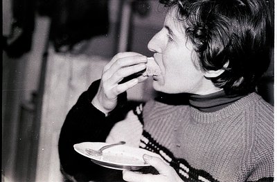Black-and-white candid shot of an individual eating at a casual indoor setting, likely mid-20th century. Person wears a patte...