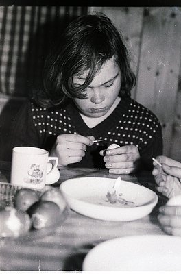 Mid-century domestic scene: Woman in patterned sweater examines food on a plate with a spoon, beside a floral-patterned mug. ...