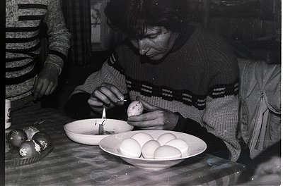 Black-and-white photograph of a woman decorating Easter eggs with a wax pen, seated at a table with a bowl of undyed eggs. Sh...