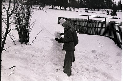 Child sculpting a snowman in a winter garden, mid-20th century. Wooden fence and bare trees frame the scene, suggesting rural...