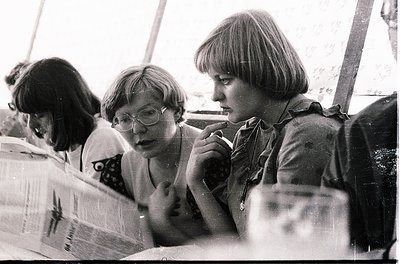 Black-and-white candid shot of three women in casual 1970s attire—short-sleeved blouses, glasses, and shoulder-length hair—en...