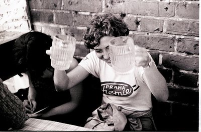 Vintage black-and-white photo of two individuals playfully holding empty glasses in a dimly lit, brick-walled room. The perso...