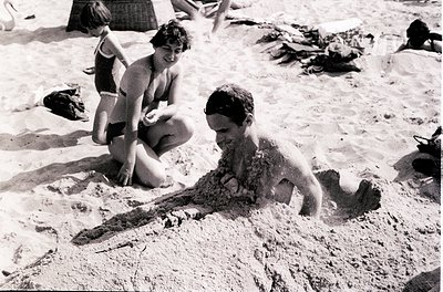 Mid-20th century beach scene: children playing in sand, one crawling while another kneels nearby. Classic 1950s–60s swimwear ...